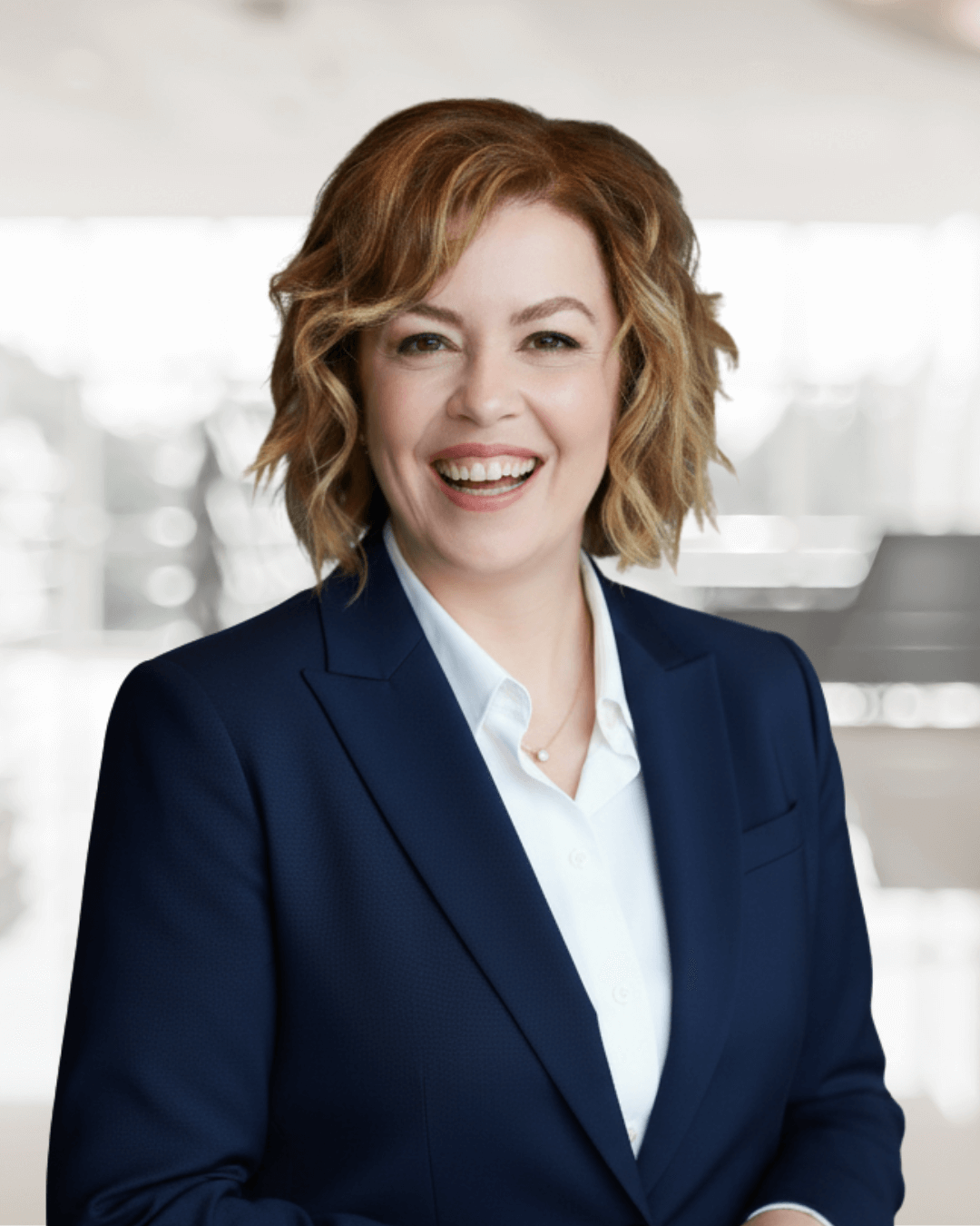 Smiling woman with wavy brown hair wearing a navy blazer and white shirt, standing in a softly blurred office setting, conveying positivity.
