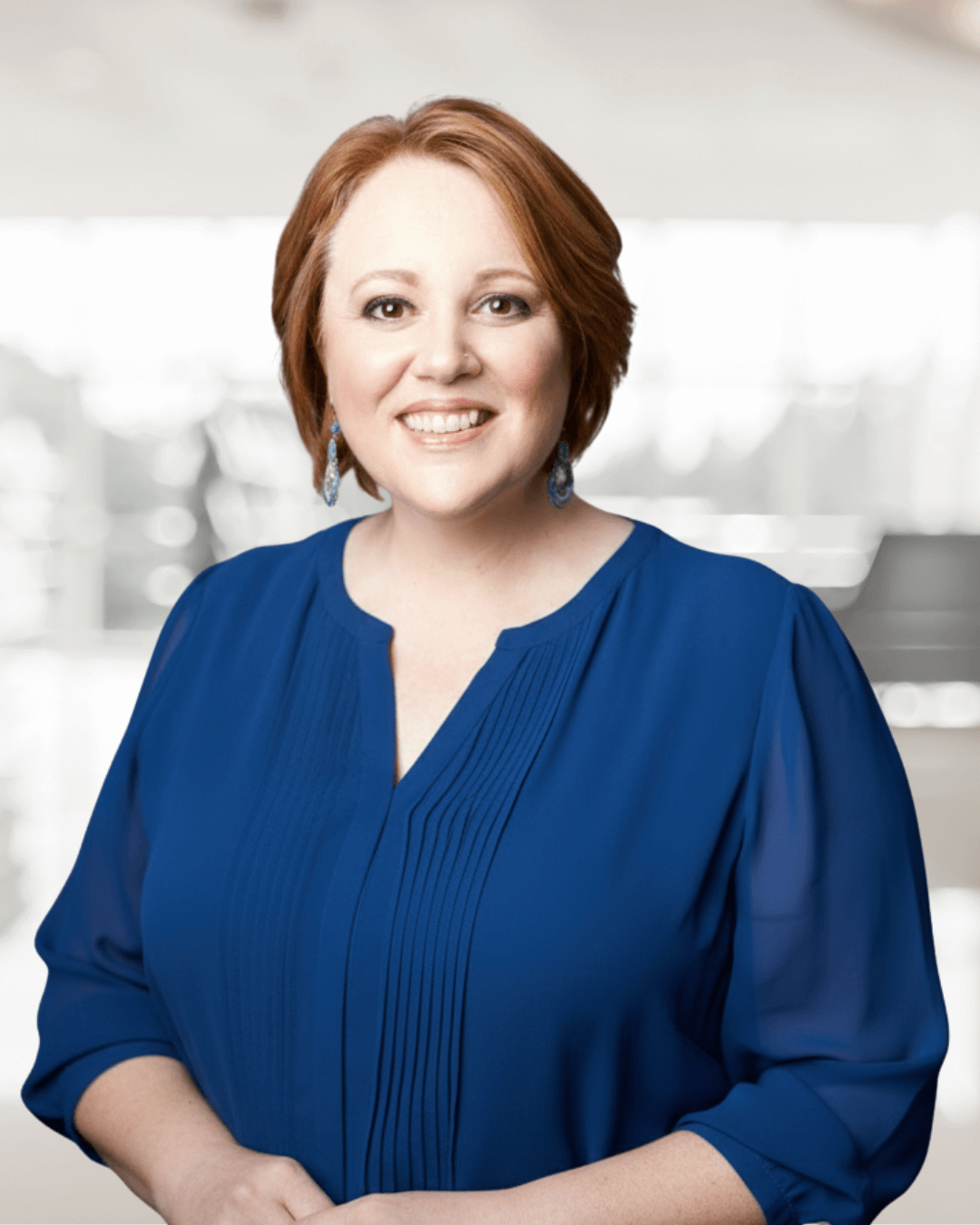 Smiling woman with short red hair wearing a blue blouse and earrings, standing in a softly blurred indoor setting, conveying a warm, professional tone.