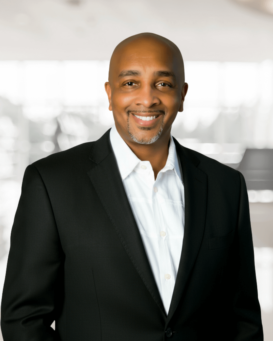 Smiling man in a black suit and white shirt stands against a softly blurred, bright background. The atmosphere is professional and approachable.