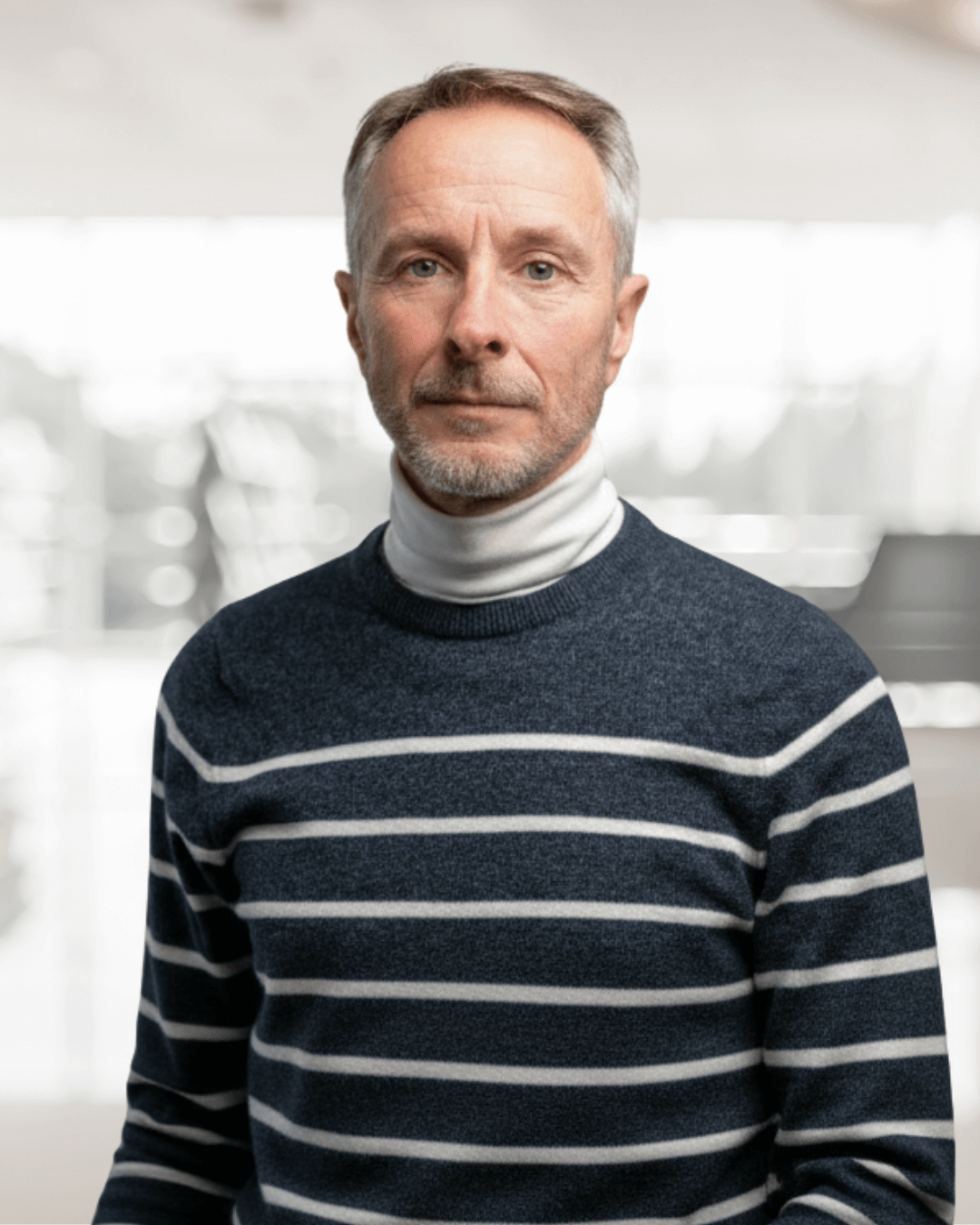 A middle-aged man with short gray hair and a beard wears a white turtleneck and a navy blue striped sweater, standing confidently indoors.