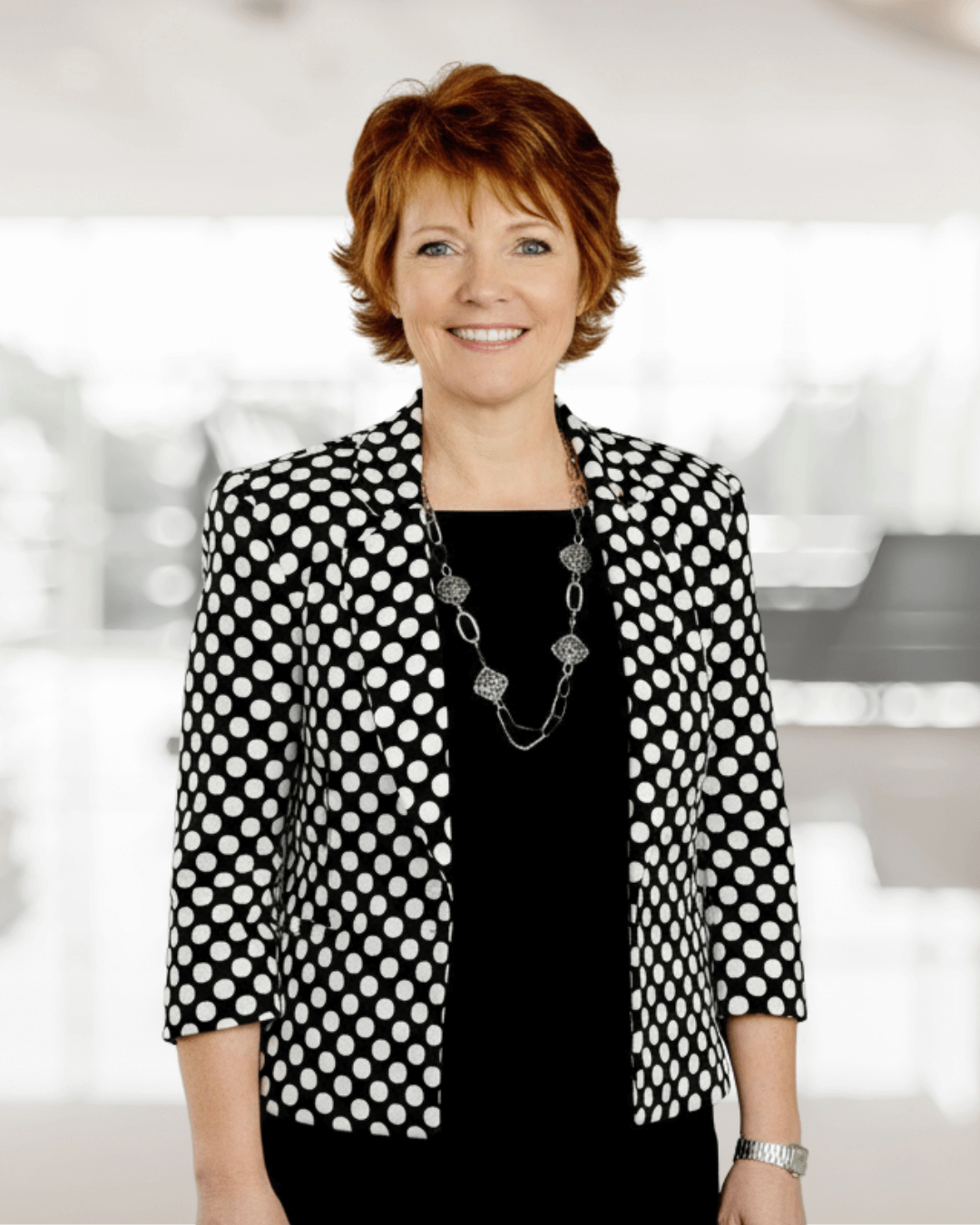 Smiling woman with short auburn hair wearing a black dress and polka dot jacket. She stands confidently in a bright, blurred office setting.