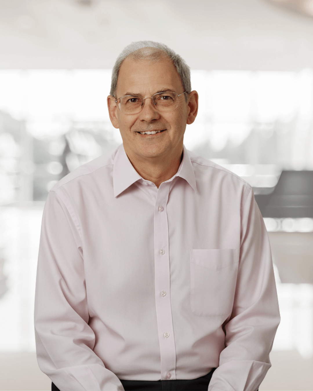 Smiling older man in glasses and a light pink shirt sits indoors, with a softly blurred background of a modern, brightly lit office space.