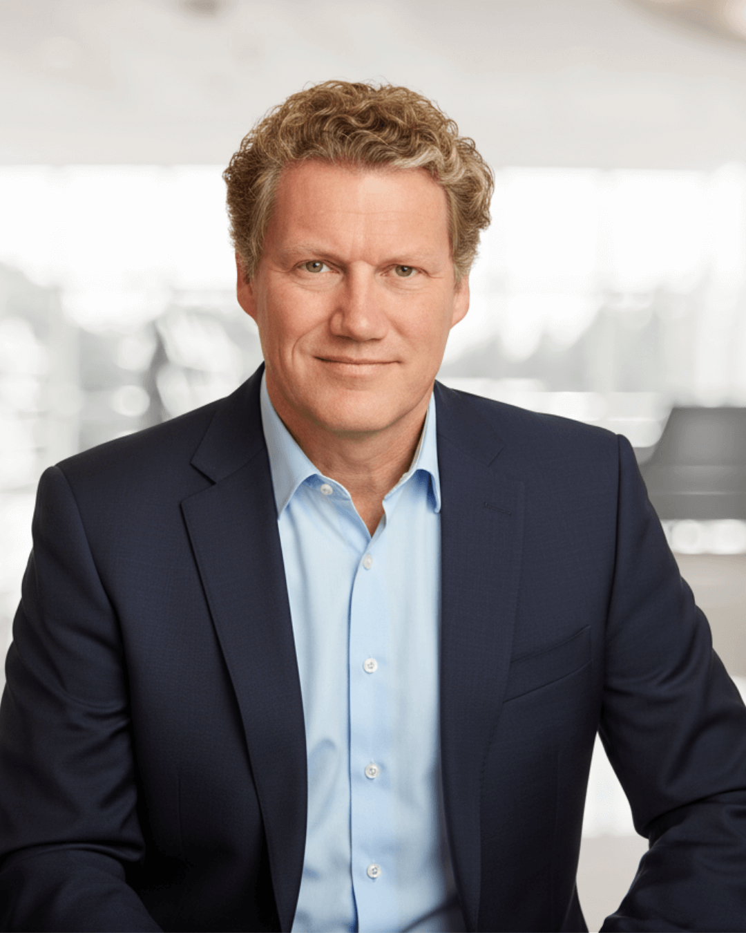A man in a blue suit and light blue shirt, with curly blond hair, smiles softly against a blurred, bright indoor background, conveying professionalism.