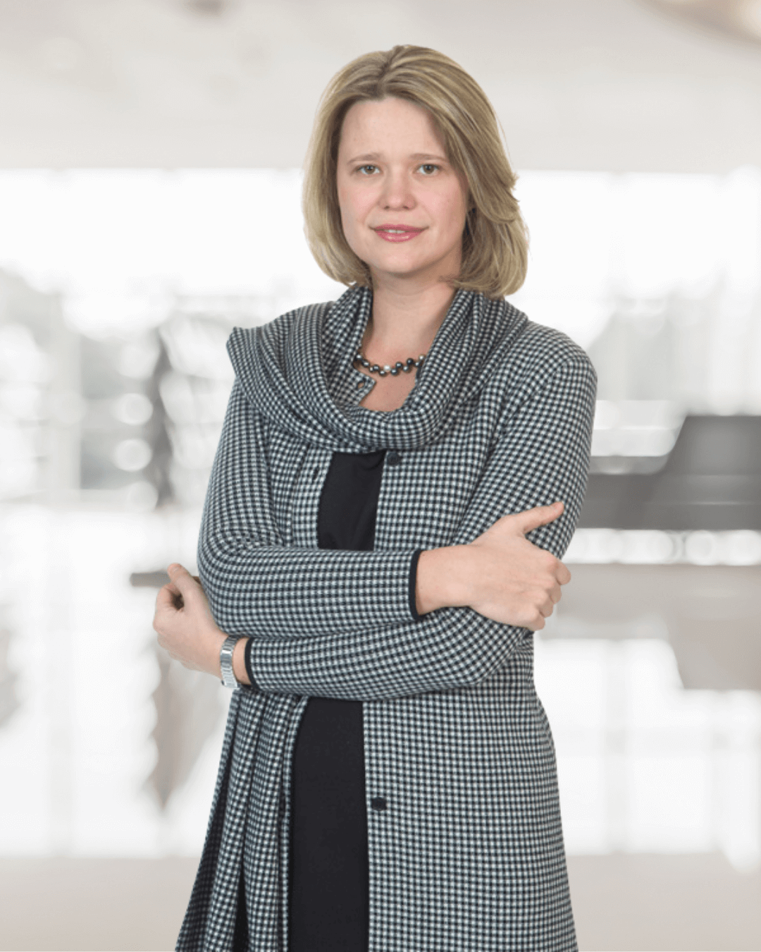 A woman with shoulder-length blonde hair is confidently posing with crossed arms. She wears a black and white checkered outfit and a necklace. The background is softly blurred, suggesting an indoor setting.