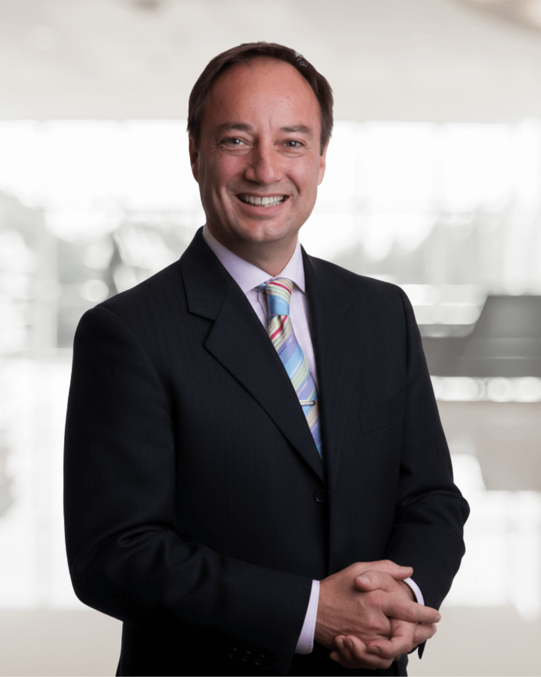 Smiling man in suit with colorful tie stands in bright, blurred office. Hands clasped, he exudes confidence and warmth.