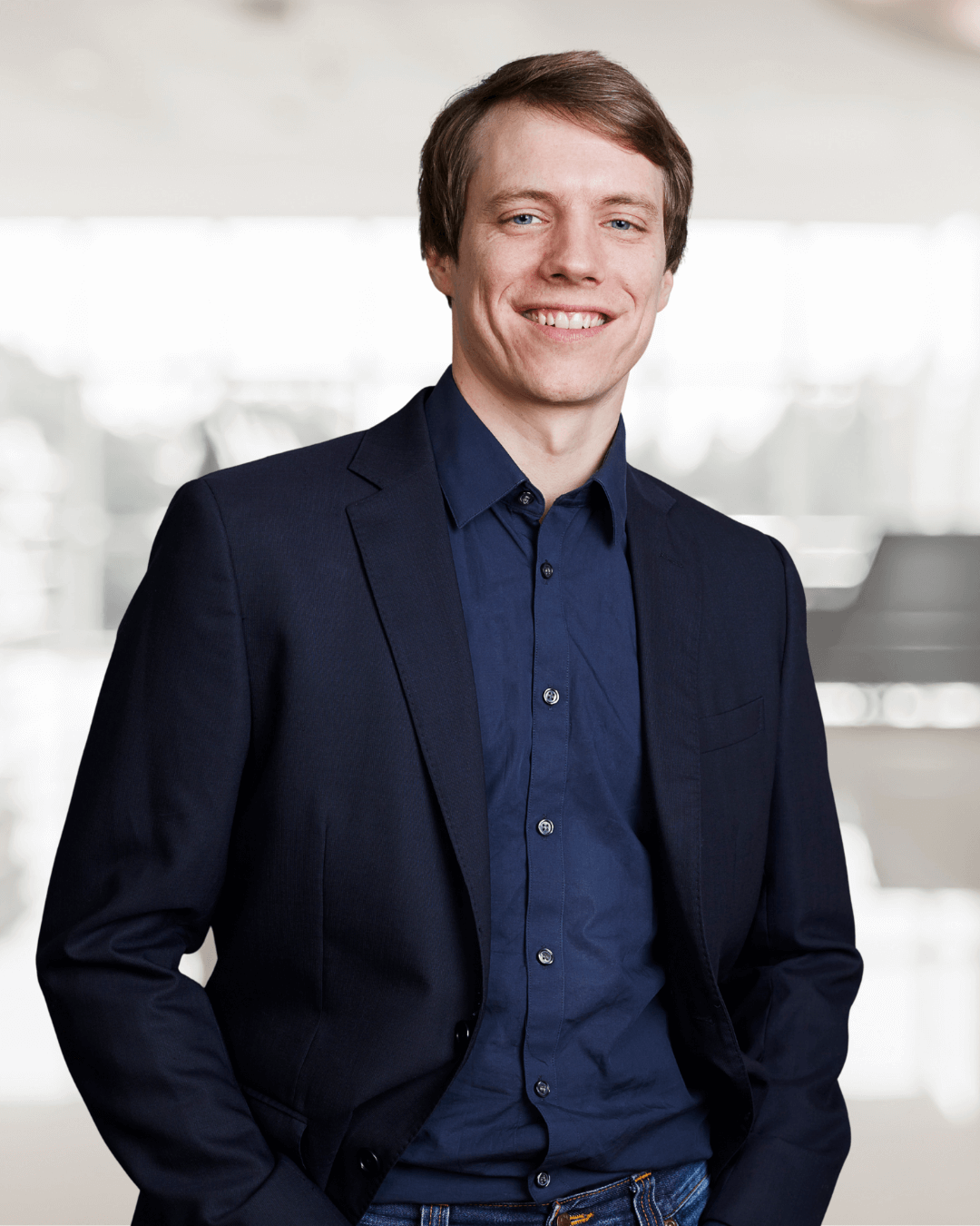Smiling man in a navy blazer and shirt stands confidently with hands in pockets. The backdrop is a softly blurred office environment, conveying professionalism.