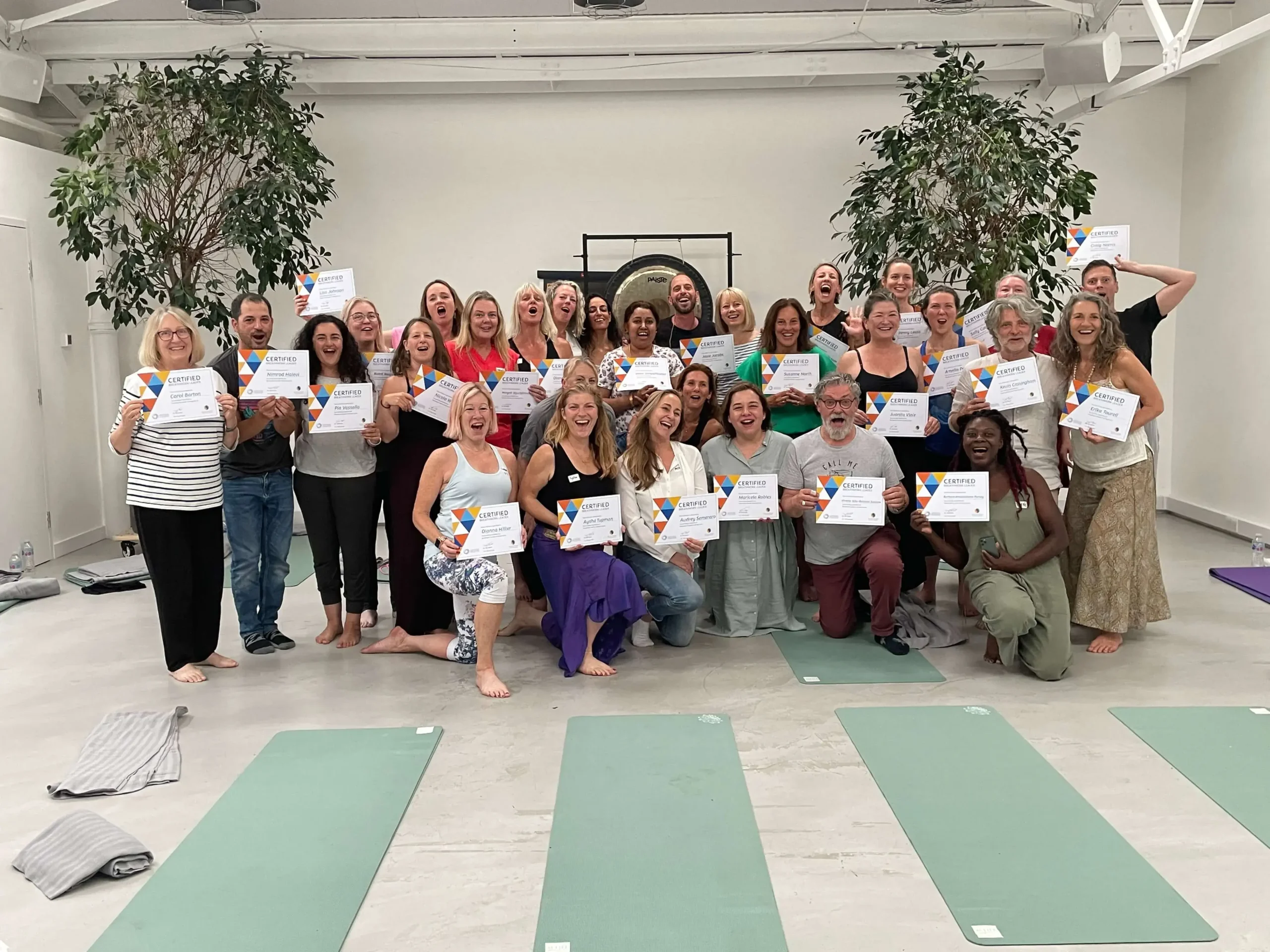 A group of people smiling, holding certificates in a bright yoga studio with mats on the floor. The atmosphere is joyful and celebratory.