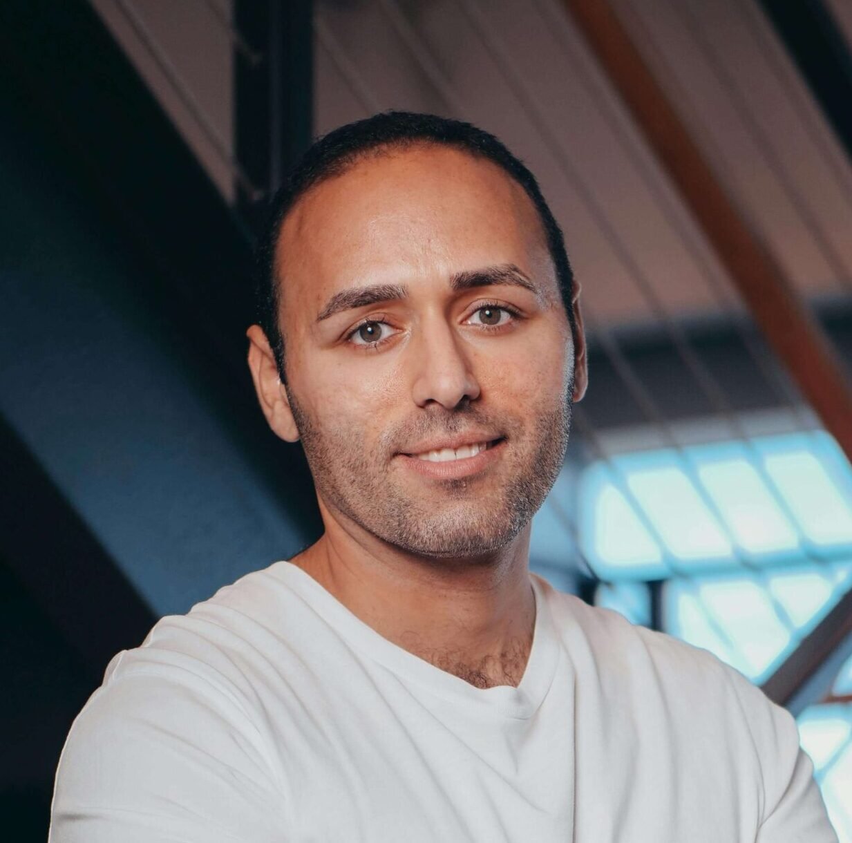 Man in a white t-shirt with arms crossed, standing in front of a modern staircase. He has a relaxed, confident expression. Natural light streams in.