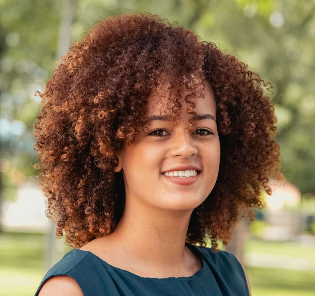 Young woman with curly hair smiling, wearing a sleeveless blue dress, stands in a sunlit park with green grass and trees in the background.