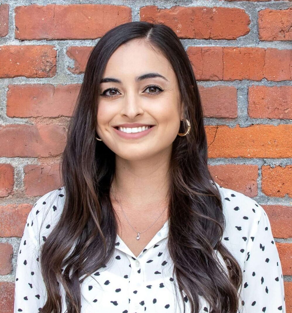 A woman with long, wavy hair smiles warmly, standing against a rustic brick wall. She wears a white polka dot blouse and black pants, exuding a professional yet approachable vibe.